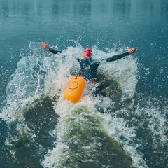 Enthusiastic swimmer in wetsuit and red cap, orange buoy trailing behind, rushes into lake water...