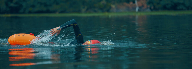 Banner. Swimmer in wetsuit and red cap swims with orange buoy in calm lake, creating splashes,...