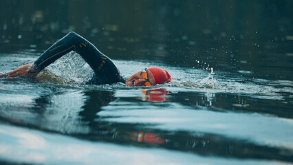 Focused swimmer in wetsuit and red cap powers through the water with orange buoy in tranquil river....