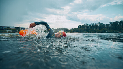 Gliding through river water, female athlete in black wetsuit and red swim cap accompanied by orange buoy. Training outdoor. Concept of sport competition, workout, recreation, healthy lifestyle.