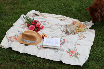 Bouquet of flowers, straw hat, summer fruit and an open book on a picnic blanket. Romantic date in a park. Summer still life photo. 