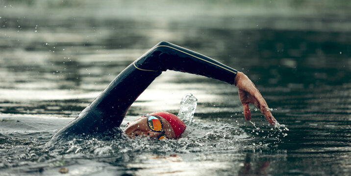 Triathlete in red swim cap and black wetsuit practices freestyle stroke, arms cutting through water, creating ripples on river's surface. Concept of sport competition, workout, recreation.