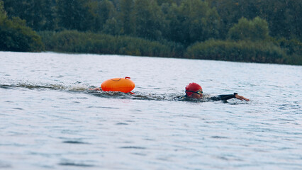 Athlete in red swim cap and black wetsuit glides through calm waters, perfecting freestyle technique, surrounded by trees. Concept of sport competition, workout, recreation, healthy lifestyle.