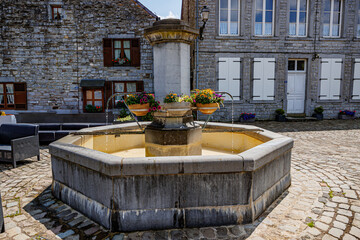 Old fountain in public square in picturesque village of Hierges, flower pot holders with flower pots, buildings with stone walls in background, sunny summer day in the Ardennes departement of France