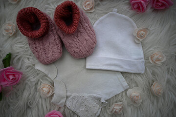 Pink knitted baby booties and a white hat lying on a white fur surrounded by pink roses