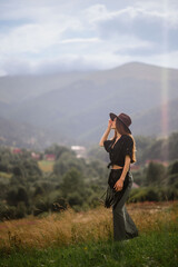 Fashionable confident woman wearing trendy hat, summer linen shirt, green maxi skirt, carrying black leather bag with fringe, walking among mountains. Outdoor full-length fashion portrait