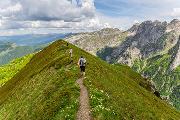 Three hikers follow a trail through a green hill next to a magnificent mountain range. 