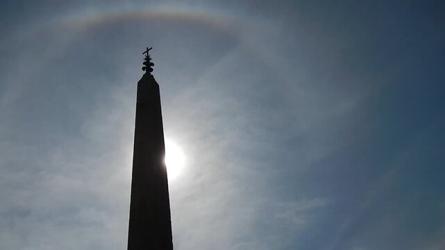 Sallustiano Obelisk with cross, ancient Roman copy of Egyptian obelisk in Rome in Italy, blue sky, sun with halo and Upper tangent arc on Cirrus clouds and birds in flight.