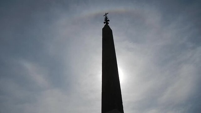 Sallustiano Obelisk with cross, ancient Roman copy of Egyptian obelisk in Rome in Italy, blue sky, sunlight with halo and Upper tangent arc on Cirrus clouds and birds in flight.