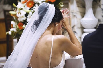 View from behind a white Caucasian woman dressed as a bride in a white dress and veil on her head, kneeling and praying in a Catholic church at the altar