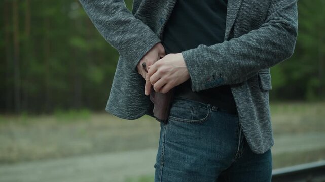 A close view of a man removing a concealed firearm under his jacket, standing beside a railway track, with blur forest view