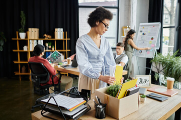 Trendy african american woman packing her items during lay off, colleagues on backdrop.