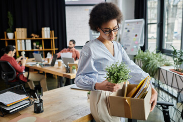 Fototapeta premium Good looking african american woman packing her items during lay off, colleagues on backdrop.