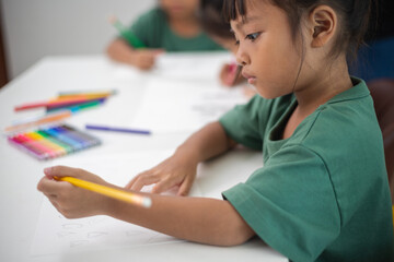 A young girl is sitting at a table with a pencil and a piece of paper in her hand. She is practicing drawing and coloring, honing her skills at home.