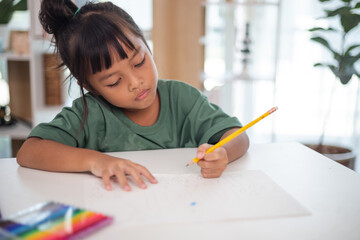 A young girl is sitting at a table with a pencil and a piece of paper in her hand. She is practicing drawing and coloring, honing her skills at home.