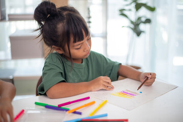 A young girl is sitting at a table with a pencil and a piece of paper in her hand. She is practicing drawing and coloring, honing her skills at home.