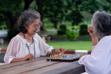 Two elderly women have gray hair and are dressed in light-colored are engaged in a friendly game of chess outdoors. They appear to be enjoying thoughtfully holding a chess piece  this older lifestyle.