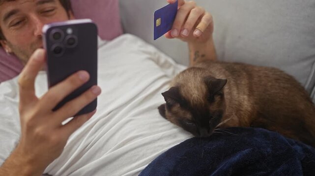 A man takes a selfie with a credit card and his siamese cat while lounging on a couch at home.