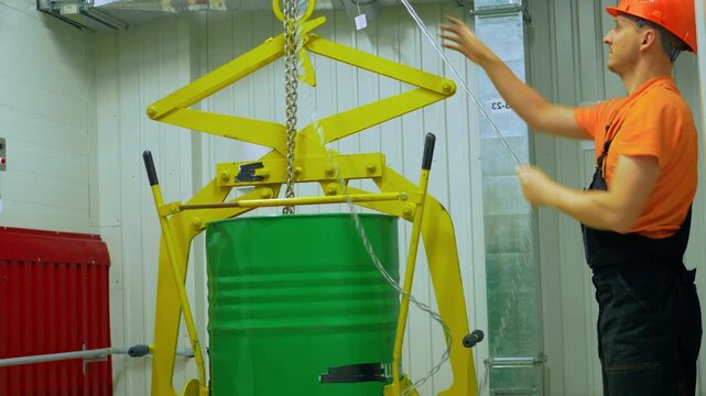 Worker lifts chemical waste barrel with chain hoist in industrial plant. Emphasizing safety protocols, protective gear. Chemical waste handling requires precision, caution. Worker ensures secure.