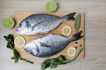 Fresh Fish with Lemon and Herbs on Cutting Board. Two fresh fish are placed on a wooden cutting...