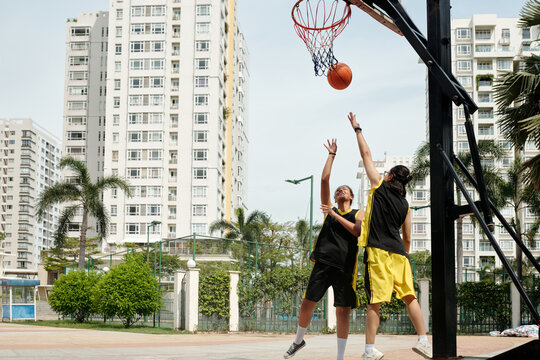 Two young men playing basketball on urban outdoor court surrounded by high-rise buildings. One player is attempting a layup while other is trying to block shot