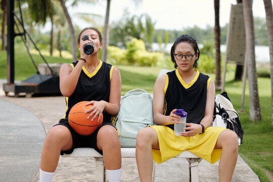 Two friends drinking water and resting after an intense basketball game on an outdoor bench by the court in the park. Both wearing basketball uniforms