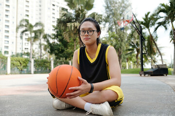 Young Asian female basketball player sitting on outdoor court in urban setting Holding orange basketball with serious expression surrounded by tall buildings and palm trees