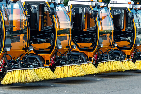 Several orange street sweepers aligned in a row, with large brushes visible in the foreground, parked on an asphalt surface