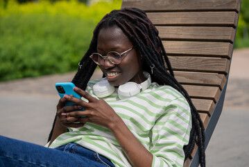Black woman with headphones relaxes on a wooden bench outdoors, enjoying her smartphone on a sunny day in a park. 