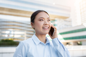 A young Asian lady professional with a friendly smile is engaged in a lively phone conversation outside. The modern architecture in the background suggests an urban environment