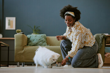 Woman kneeling and feeding her small white dog indoors while wearing white blouse and blue jeans sitting by stylish furnishings in comfortable home