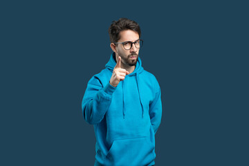 A young man in a blue hoodie stands confidently against a plain blue backdrop, raising his index finger as if emphasizing a point. His expression is serious, showcasing determination and focus