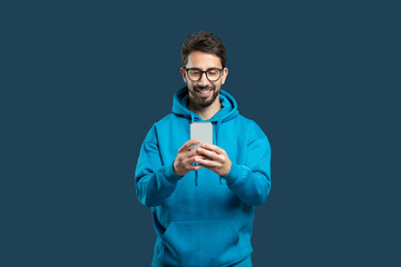 A young man wearing a bright blue hoodie stands against a solid background, happily engaging with his smartphone. His glasses reflect excitement as he interacts with the device