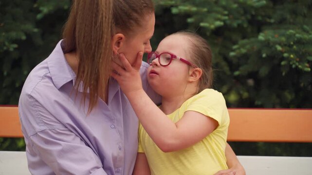 Mother and her young daughter with Down syndrome sit on a colorful bench. Daughter gives her mother a kiss on cheek, reflecting tender and affectionate moments of nurturing child with special needs