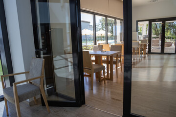 Contemporary wooden chairs and tables in the living room of a restaurant. Large windows, grass outside, umbrellas and pool.