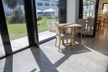 Contemporary wooden chairs and tables in the living room of a restaurant. Large windows, grass outside, umbrellas and pool.