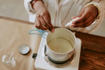 Hands carefully pouring liquid into container while standing at workspace. Surroundings include various crafting tools, hinting at meticulous, creative process