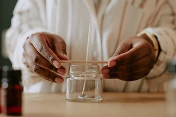 Hands crafting handmade candle in glass jar with wooden wick, surrounded by tools and ingredients for candle making. Process involves precise control and delicate handling