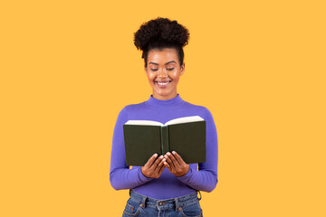 A woman stands smiling while holding a closed book, wearing a purple long-sleeve top and jeans. The...