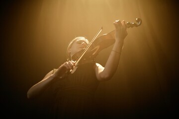 Low angle shot of female classical music artist sensually playing violin illuminated by light from...