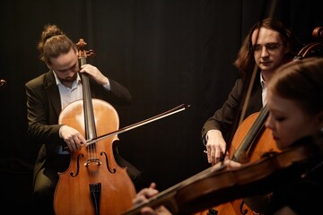 Three symphonic orchestra musicians playing cellos and violins while sitting in circle performing...