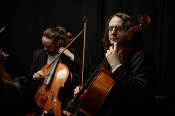 Concentrated male cello player in glasses performing classical music with string quartet in studio with dramatic top light against black background