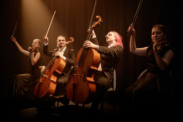 Chamber quartet of string musicians sitting in row on stage with project light raising bows while...