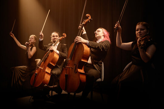 Chamber quartet of string musicians sitting in row on stage with project light raising bows while thanking public at end of performance