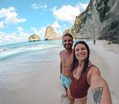 A man and woman are posing for a picture on a beach. The woman is wearing a red tank top and the man is wearing a blue swim trunks. Scene is happy and relaxed