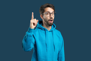 A young man wearing a blue hoodie is pointing upward with his right index finger while looking...