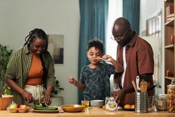 Cute little boy with closed eyes tasting food from wooden spatula while standing by kitchen table between his parent preparing breakfast