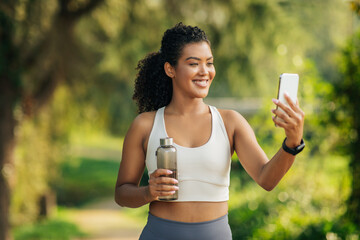 A cheerful young woman is enjoying her post-workout routine in a lush park. She holds a water bottle in one hand while capturing a selfie with her smartphone.