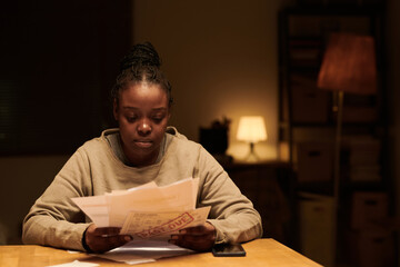 Young unhappy woman in sweatshirt holding stack of financial bills with past due stamps and looking...