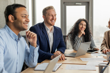 Four smiling colleagues are sitting around a table, engaged in a conversation during a meeting. The man on the left is listening intently, while the man in the middle is speaking